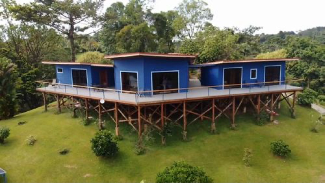 Aerial view of the blue main house on stilts, its long shared deck running the full length of the building above the green hillside